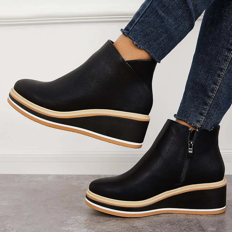 Side view of black wedge boots with zipper, worn with jeans, against a white wall and gray floor.