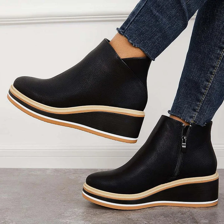 Side view of black wedge boots with zipper, worn with jeans, against a white wall and gray floor.