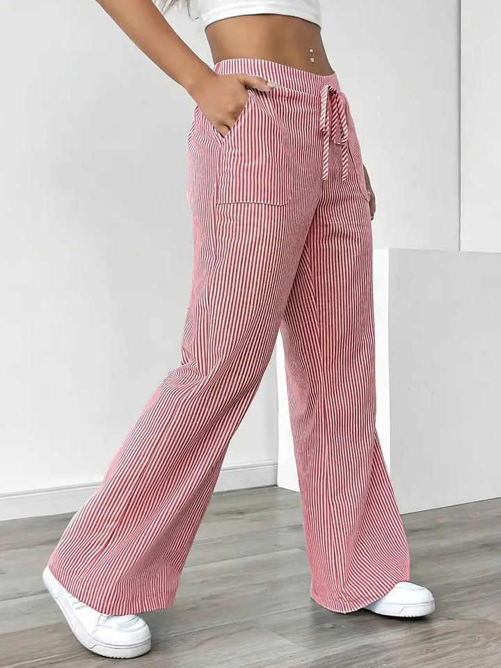 Side view of red and white striped pants with drawstring, pockets, modeled indoors, paired with white sneakers.