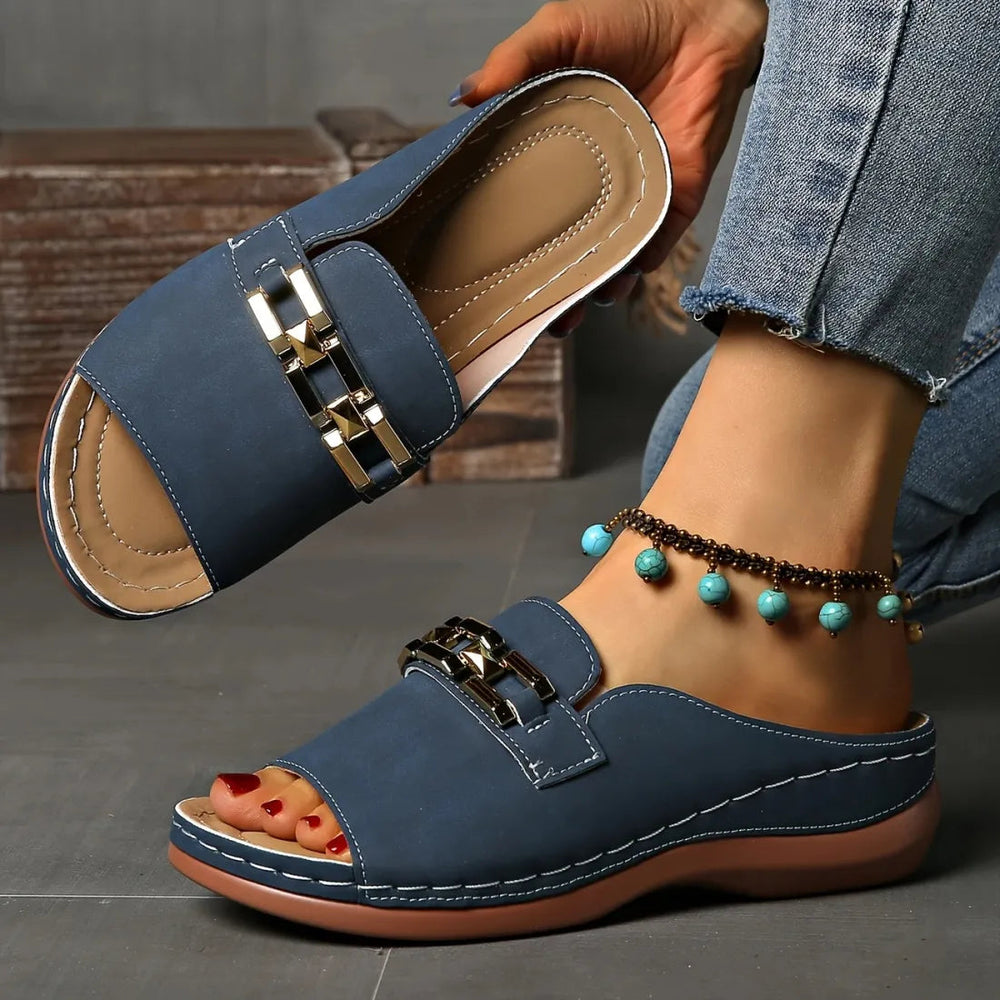 Blue elegant comfort sandals with gold accents, worn with anklet, showcased in an indoor setting on a grey tiled floor.