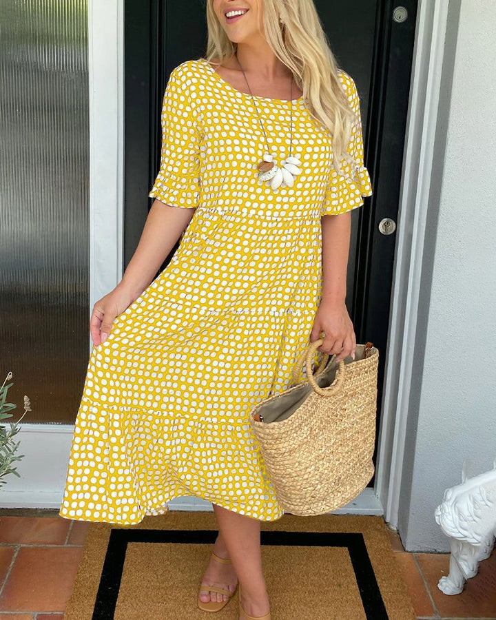 Woman in yellow and white polka dot summer dress, holding a straw tote, standing by a door.