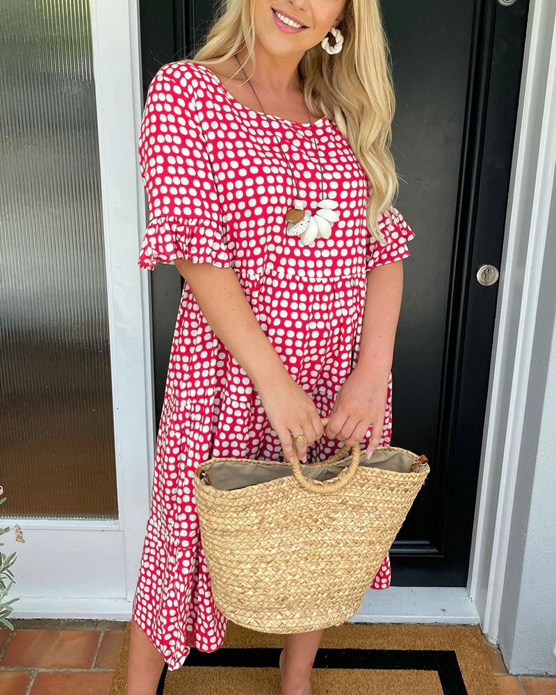 Woman in red and white polka dot summer dress, holding a straw tote, standing by a door.