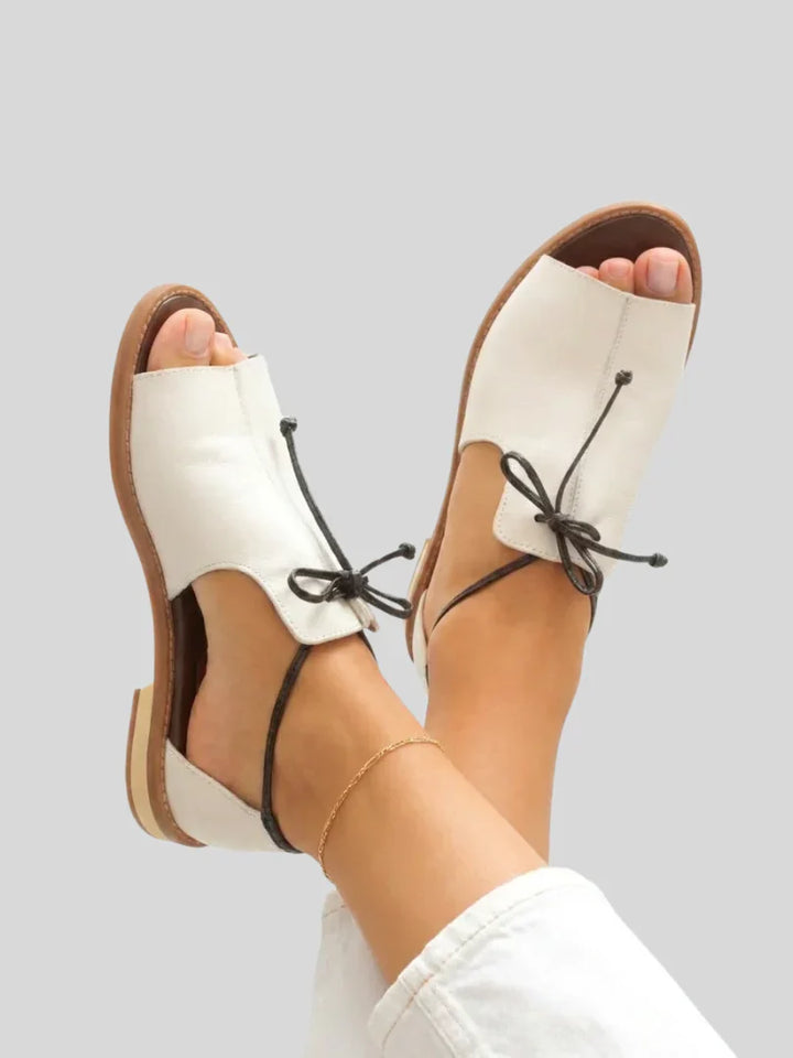 White leather sandals with black bow on feet, gray background.