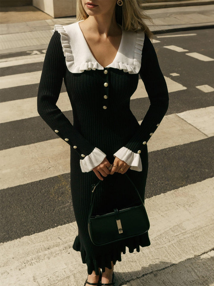 Model wearing a black knit dress with white ruffled collar and cuffs, pearl buttons, paired with a black handbag, standing on a crosswalk.