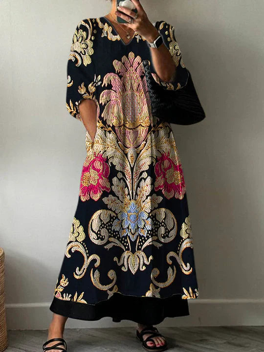 V-neck dress with intricate floral patterns in black, red, and gold, featuring pockets. Indoor setting with a light wall.