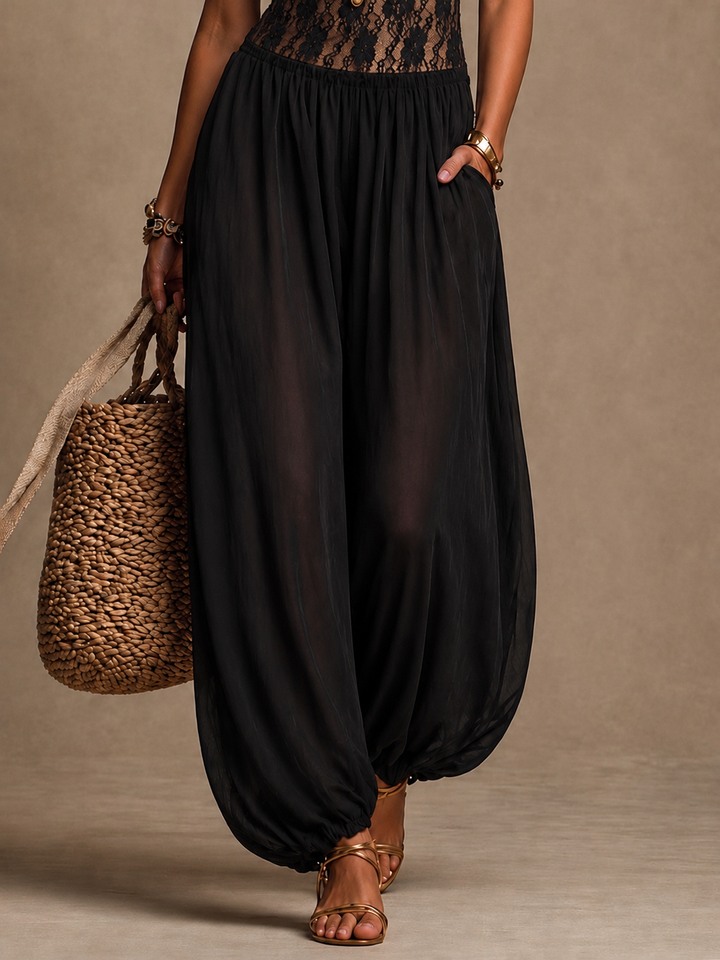 Close-up of black flowing pants of halter jumpsuit, with a straw bag and sandals, in studio setting.