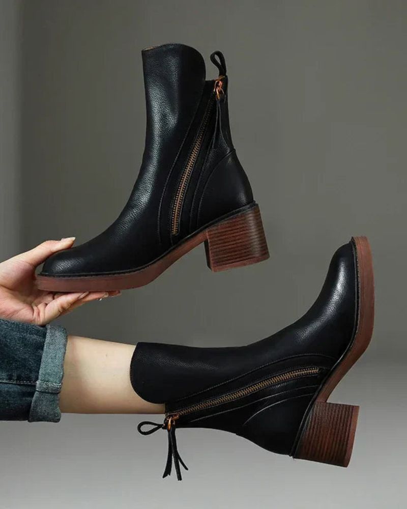 Hands holding black leather boots with zippers against a plain backdrop, showcasing design details.