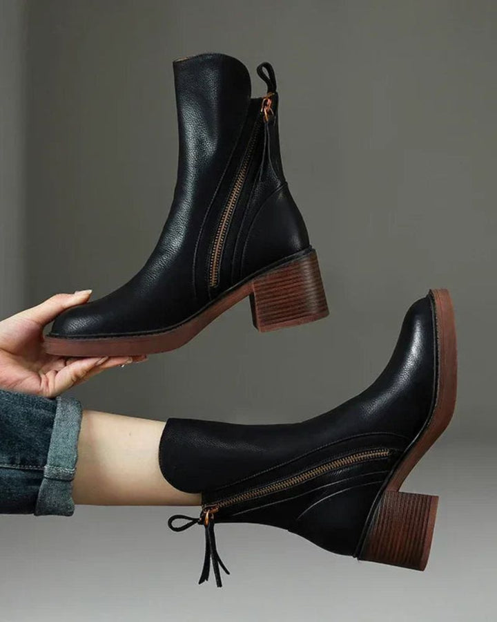 Hands holding black ankle boots with brown heels against a gray background.
