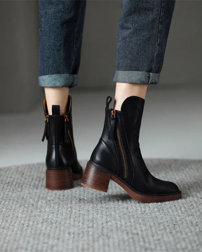 Back view of black leather boots with zippers, worn with jeans on a textured carpet in an indoor setting.