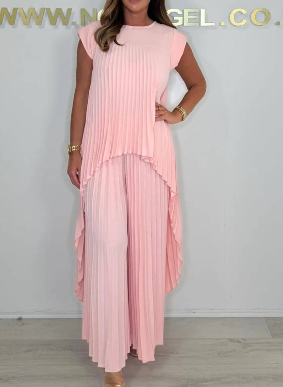 Woman in a pink pleated midi top and wide-leg pants set, posed in a bright studio with a plain wall and floor.