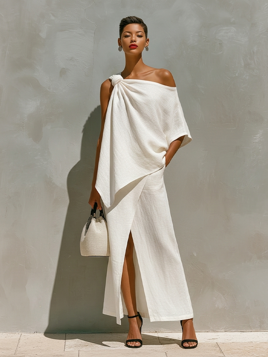 Full view of a model in a flowing white one-shoulder outfit with a slit, holding a handbag, set against a textured neutral wall.