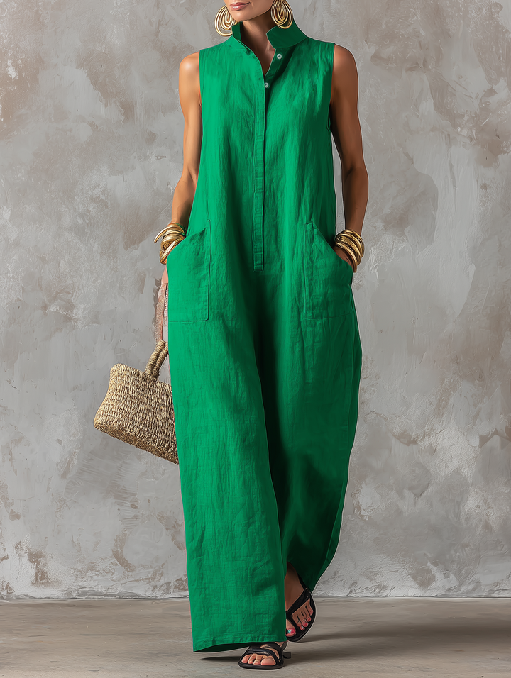 Front view of model in a green sleeveless jumpsuit with wide legs and pockets, against a textured studio background, holding a straw bag.
