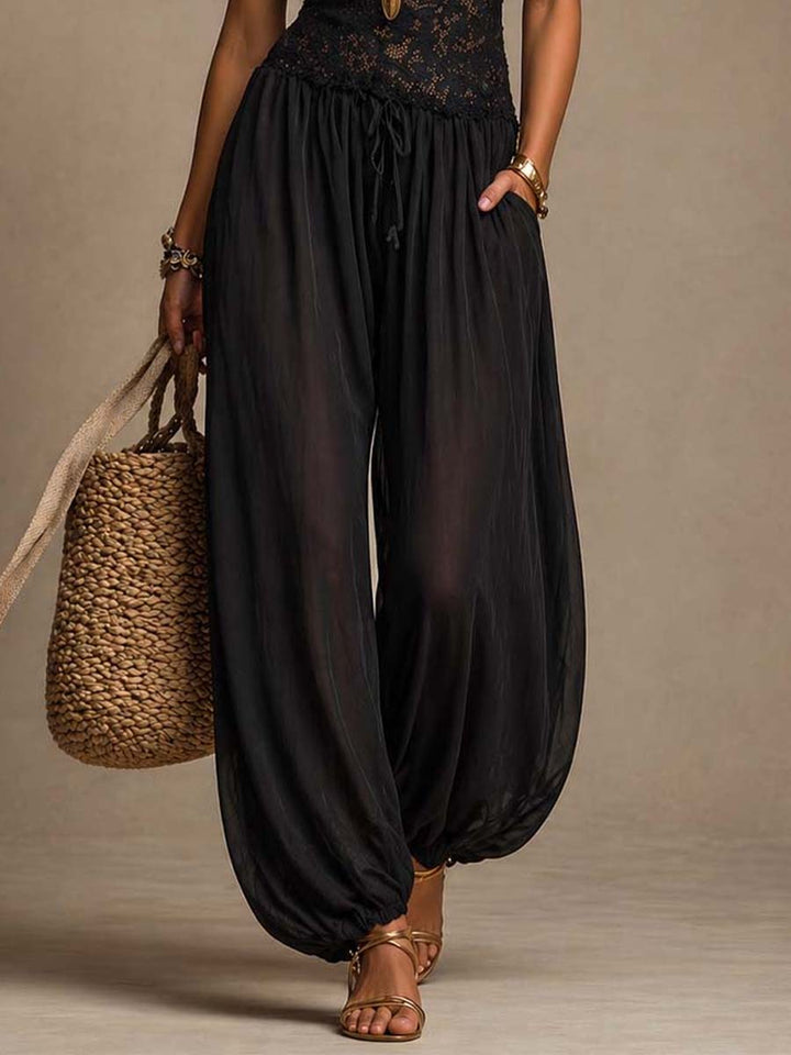 Lower view of black jumpsuit with flowy pants, accessorized with a woven bag and sandals in a studio setup.