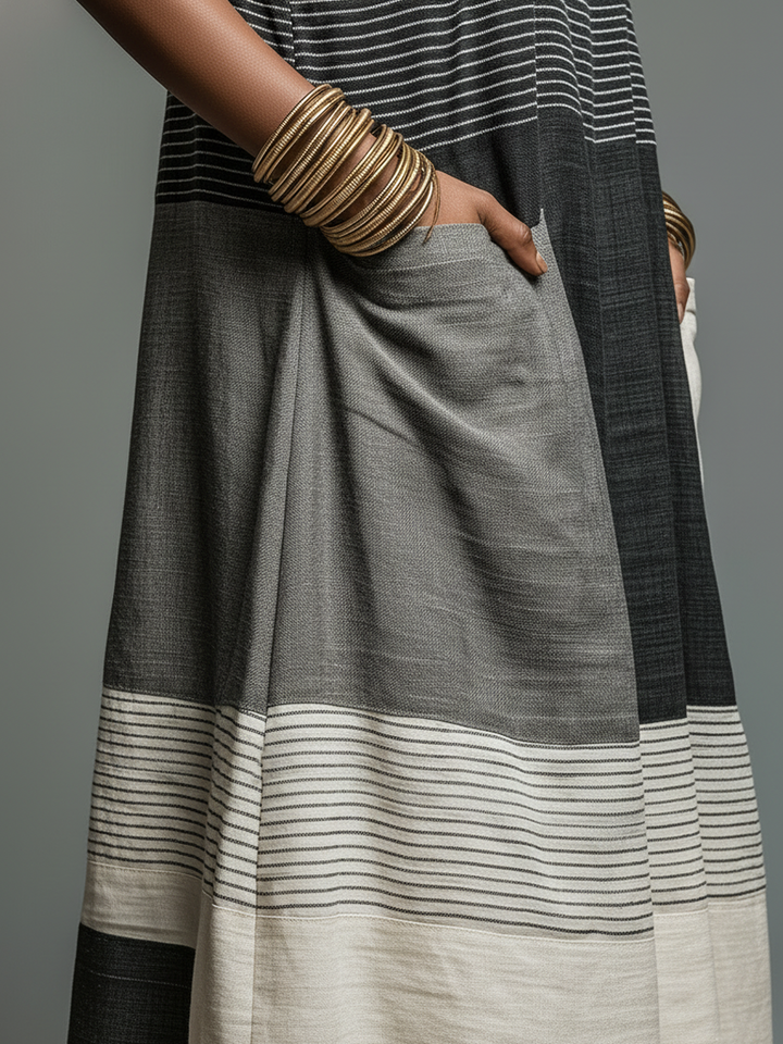 Close-up of maxi dress showing black, gray, and cream stripes and a hand in the pocket, styled with bangles, studio background.