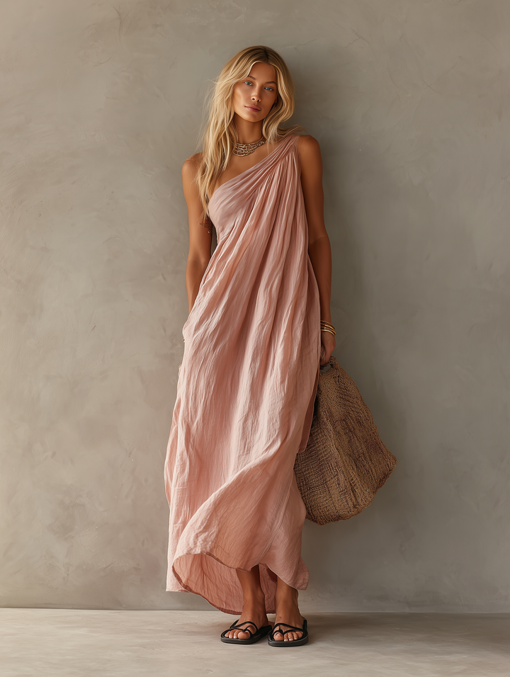 Pink maxi dress with flowing design styled with sandals and a woven bag against an indoor wall.