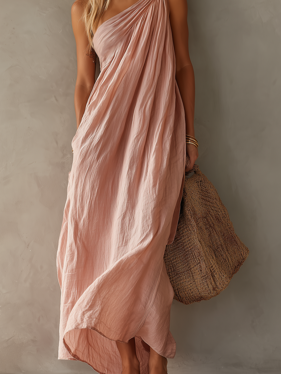 Close-up of flowing pink maxi dress with textured folds and a woven bag against a neutral wall.