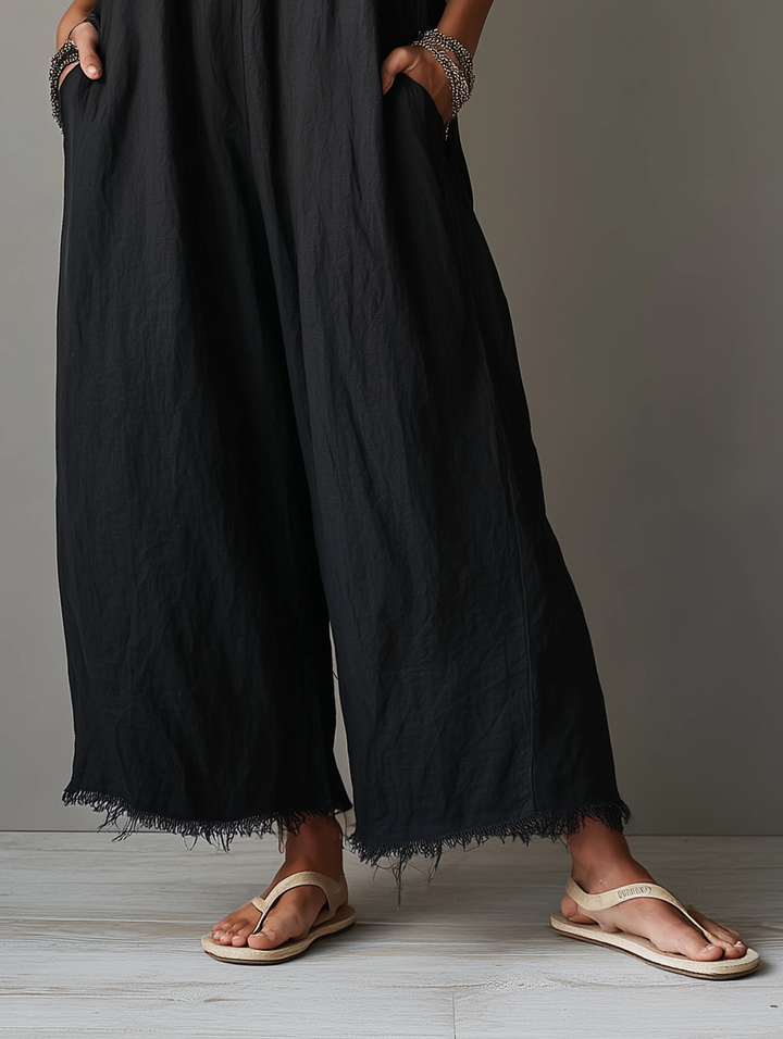 Lower half of a black jumpsuit with fringed hem, paired with beige sandals, in a studio setting.