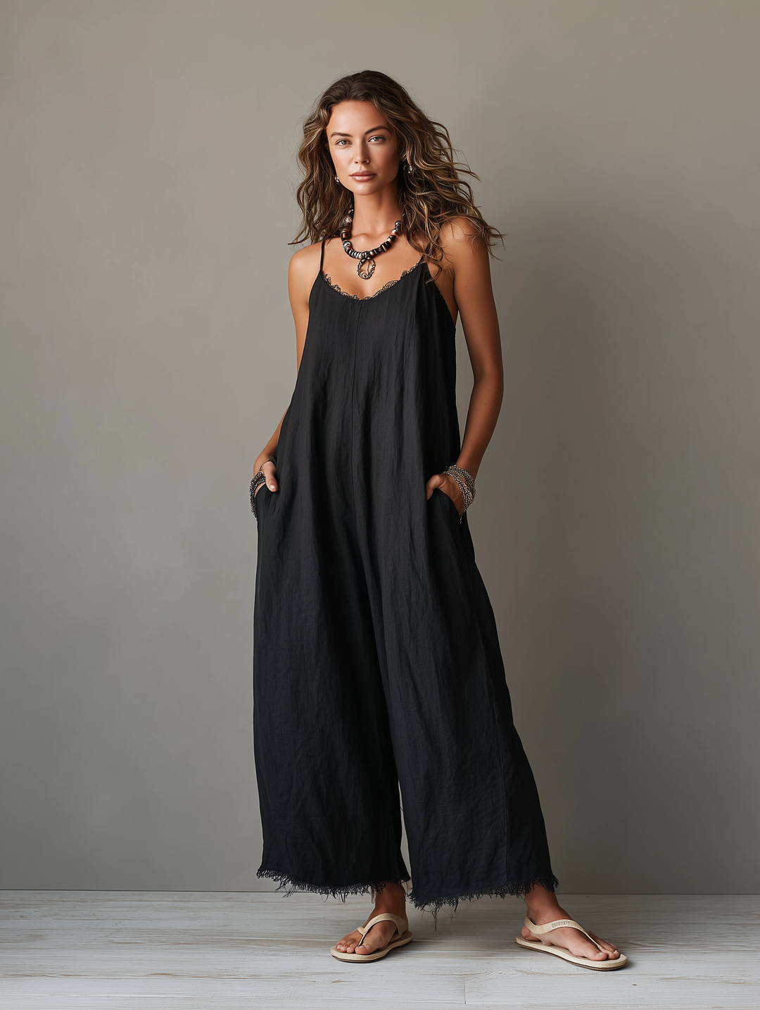 Full view of a black jumpsuit with spaghetti straps and fringed hem, model posing in sandals, studio background.