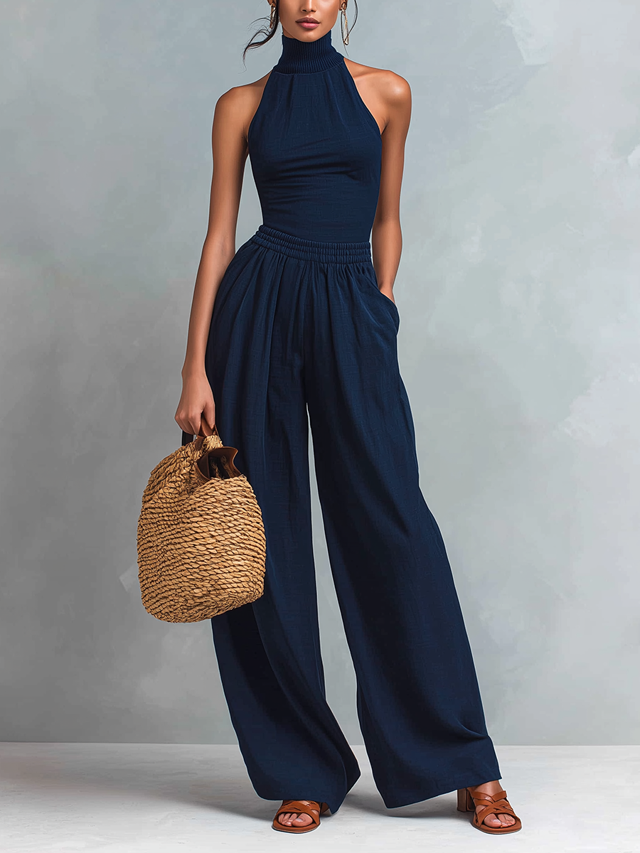 Woman wearing a dark blue halter-neck jumpsuit with wide legs, holding a woven bag in a studio setting.