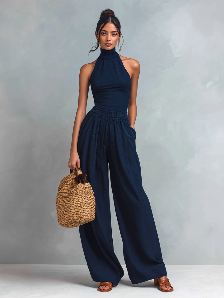 Full view of woman in elegant dark blue jumpsuit and sandals, holding a woven bag in a studio.