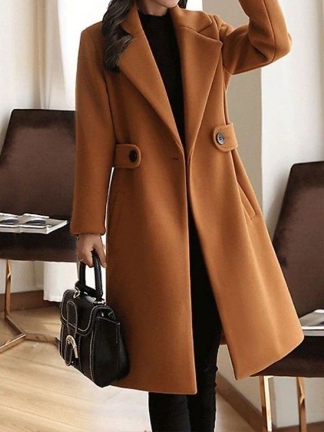 Elegant brown coat with wide lapels and button details, worn in a stylish indoor setting. Model holds a black handbag.