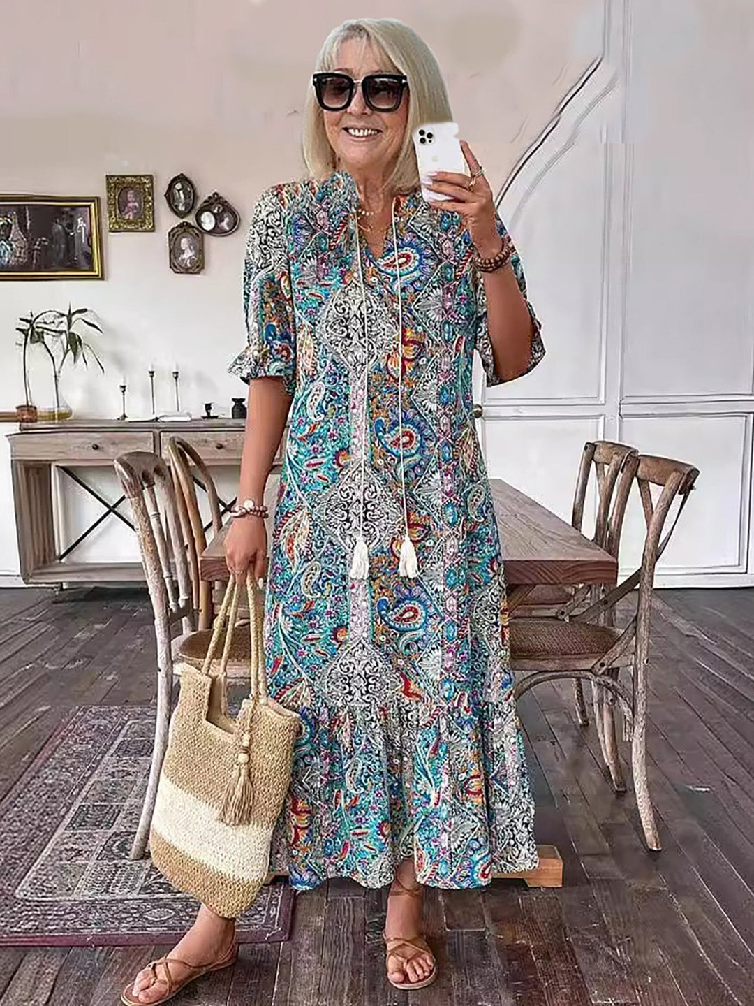 Woman in colorful boho dress, holding straw bag, indoor rustic setting with wooden furniture and framed artworks.