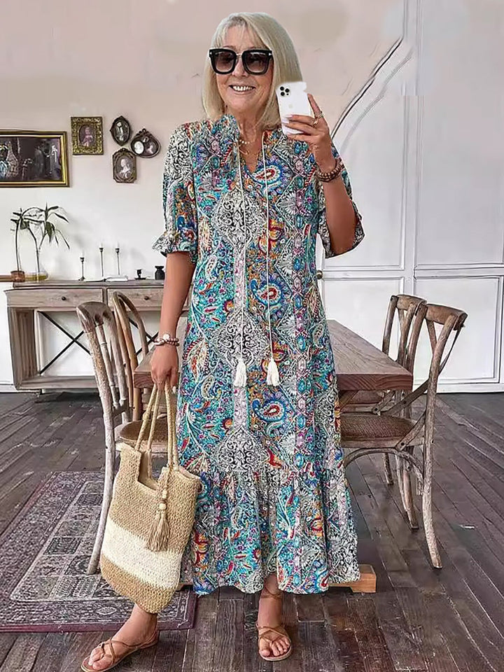 Woman in colorful boho dress, holding straw bag, indoor rustic setting with wooden furniture and framed artworks.