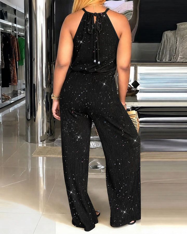 Back view of a model wearing a black glitter jumpsuit with tie closure. Indoor showroom with polished floors and garments displayed.