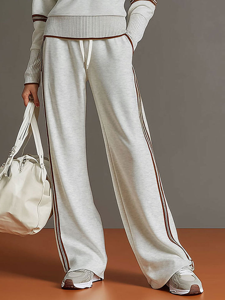 Close-up of light gray lounge pants with contrast stripes, paired with white sneakers and a bag in a studio setting.