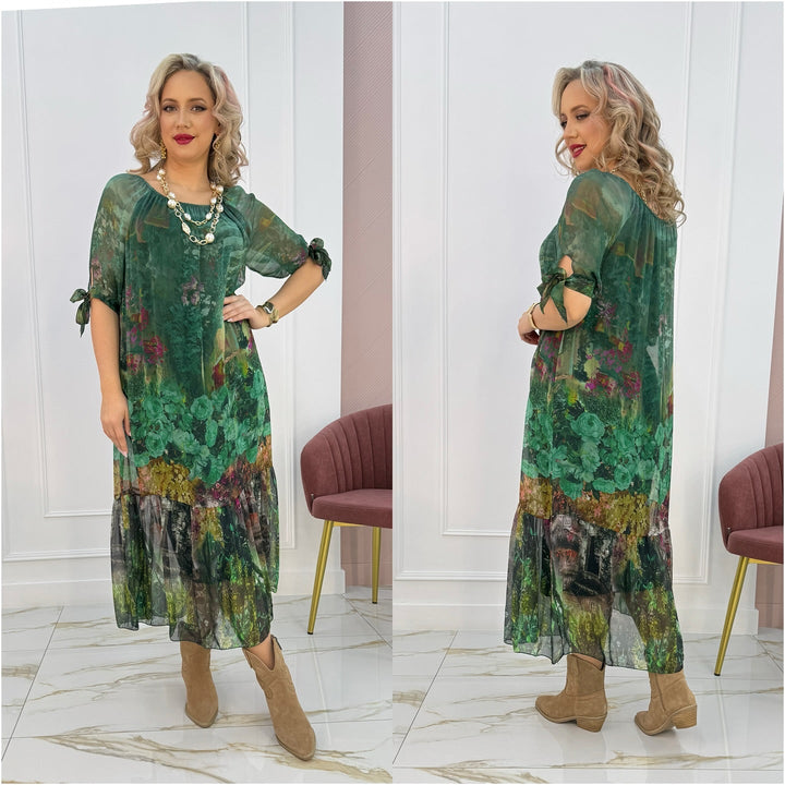 Model in green floral maxi dress with sheer overlay, standing indoors on marble floor with pink chair.
