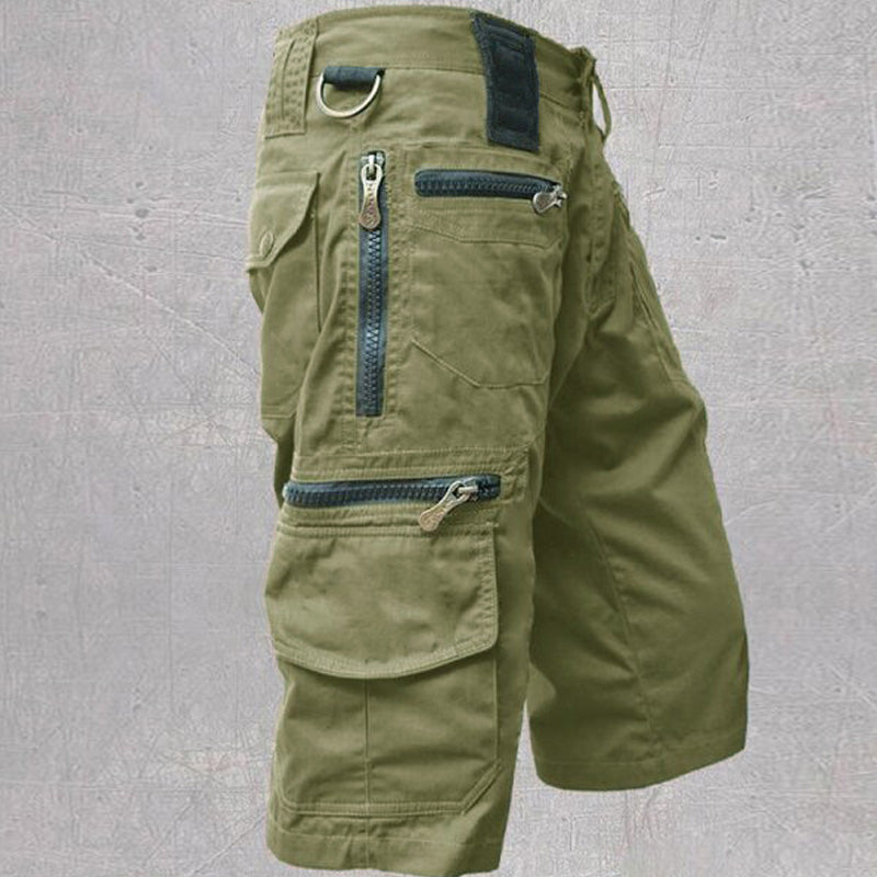 Green tactical cargo shorts showcasing zipper pockets and D-ring, contrasted with a textured gray backdrop.