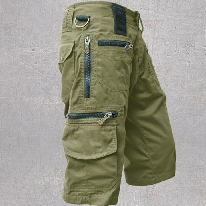 Green tactical cargo shorts showcasing zipper pockets and D-ring, contrasted with a textured gray backdrop.