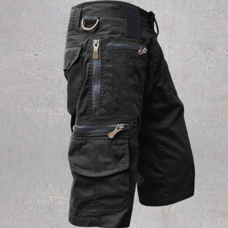 Black tactical cargo shorts featuring zippered pockets and a D-ring, set against a textured gray background.