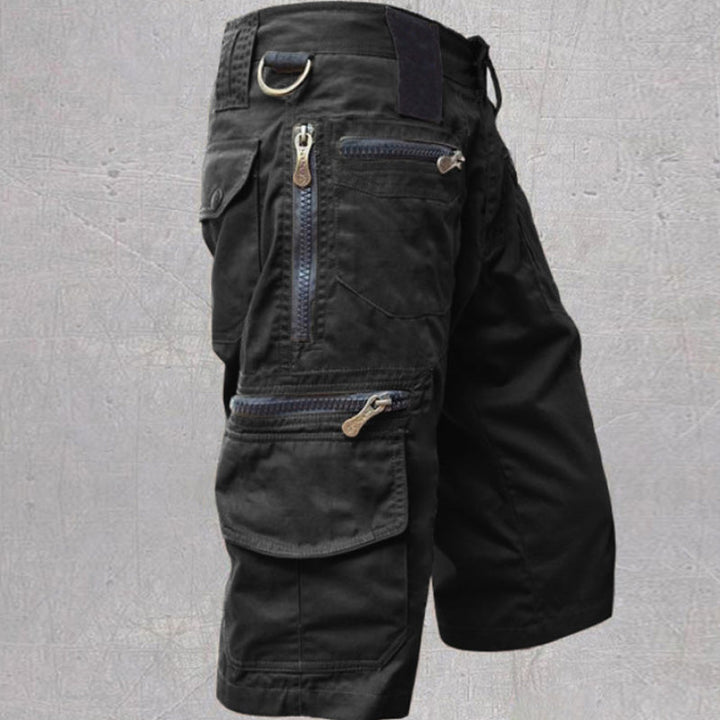 Black tactical cargo shorts featuring zippered pockets and a D-ring, set against a textured gray background.