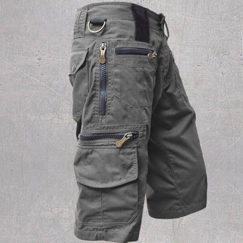 Gray tactical cargo shorts with multiple zipper pockets and D-ring, displayed against a textured gray background.