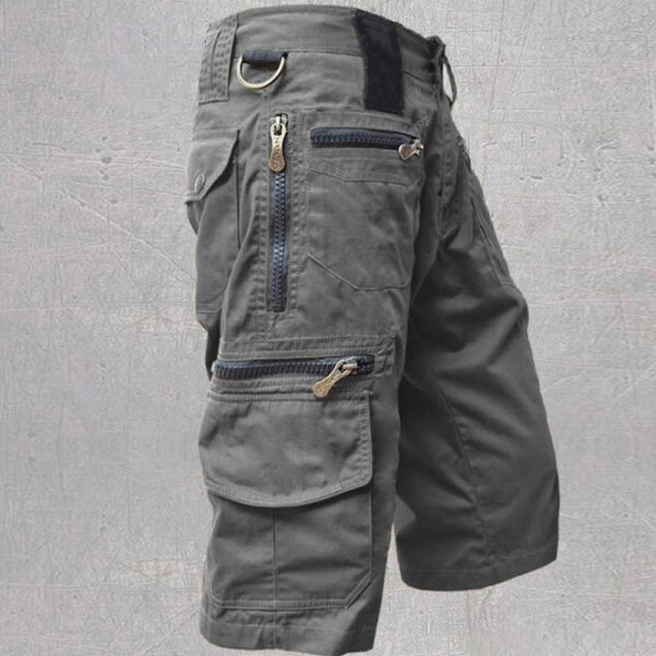 Gray tactical cargo shorts with multiple zipper pockets and D-ring, displayed against a textured gray background.