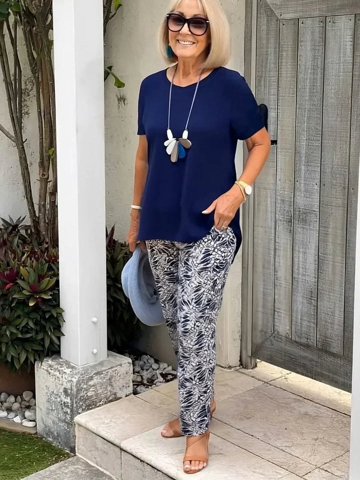 Woman in a navy loose two-piece set with abstract patterned pants, accessorized with sunglasses and a statement necklace, holding a blue hat in an outdoor setting.