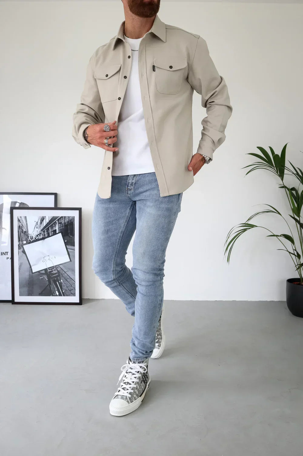 Light beige overshirt with white t-shirt and jeans, standing indoors beside frames and plant.