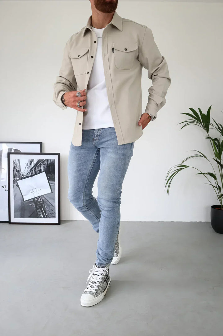 Light beige overshirt with white t-shirt and jeans, standing indoors beside frames and plant.