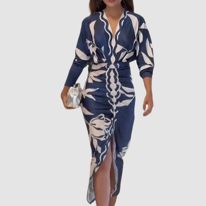 Elegant V-neck dress with bold white patterns on navy, long sleeves, and front slit. Model holding a silver clutch in a studio setting.