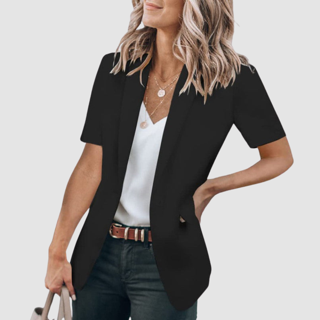 Black short-sleeve blazer worn by person with wavy hair, white top, and jeans; holding a beige handbag against a plain background.