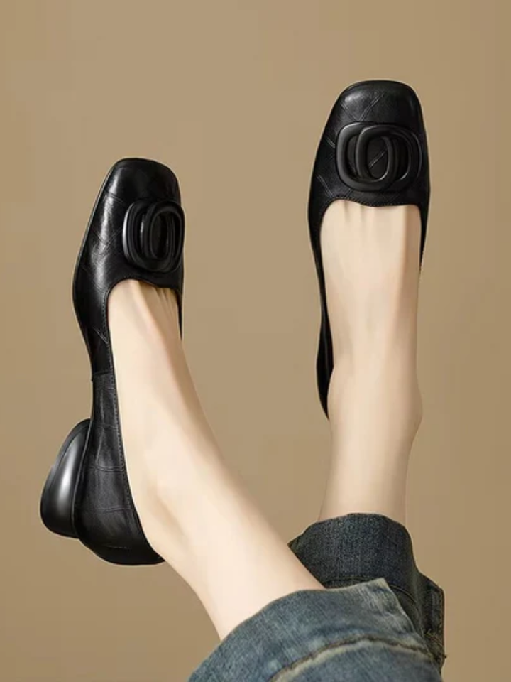 Black leather flats with ornate buckle, paired with denim, on a beige background.