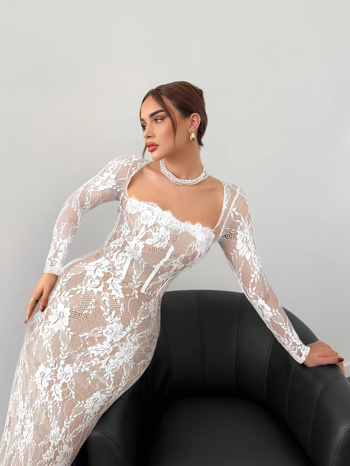 Model leaning on a black chair, wearing a detailed white lace dress with a flattering neckline in a studio environment.