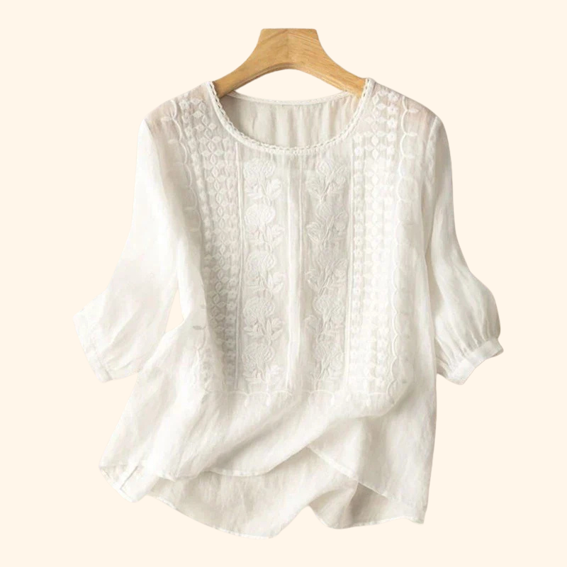 White elegant soft blouse with intricate embroidery, displayed on a wooden hanger against a beige background.