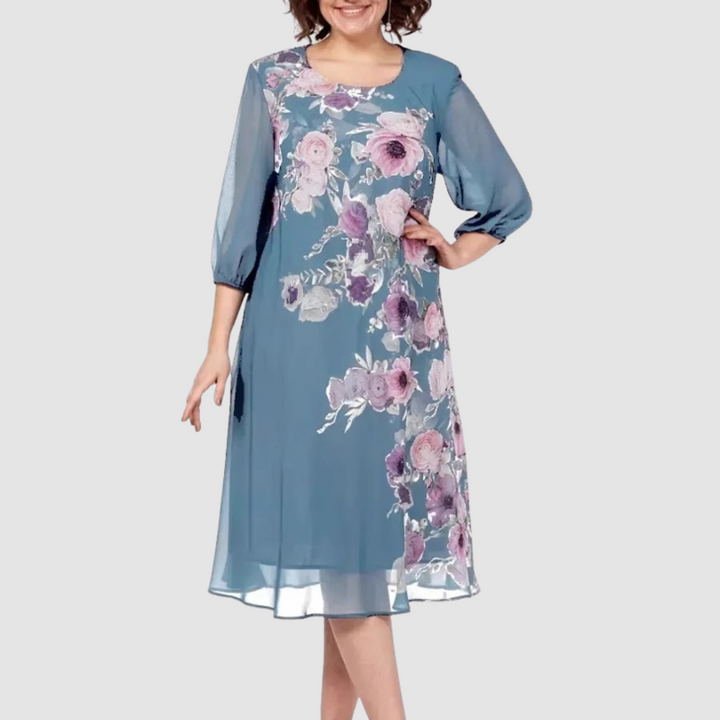 Woman in a teal floral dress with pink flowers, standing in a studio with a light background.