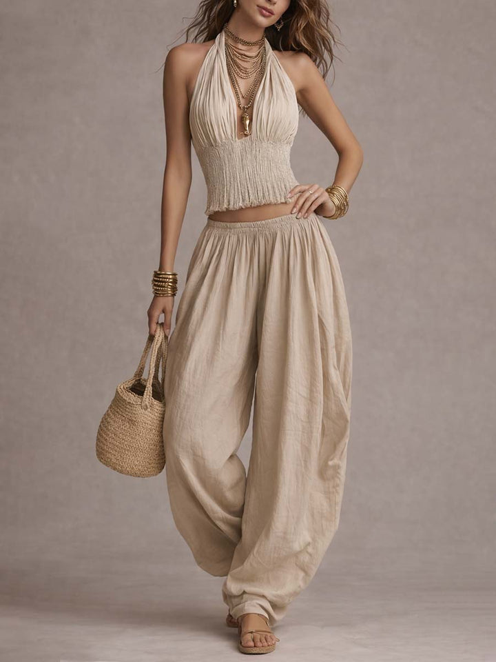 Beige two-piece set with a textured top and flowing pants, accessorized with gold bangles and a basket bag. Studio background.