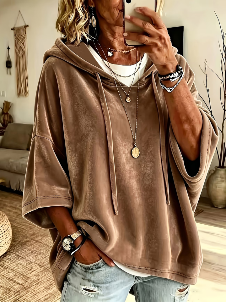Taupe oversized velour hoodie with wide sleeves and drawstrings, worn over layered necklaces in a cozy, boho-styled living room.