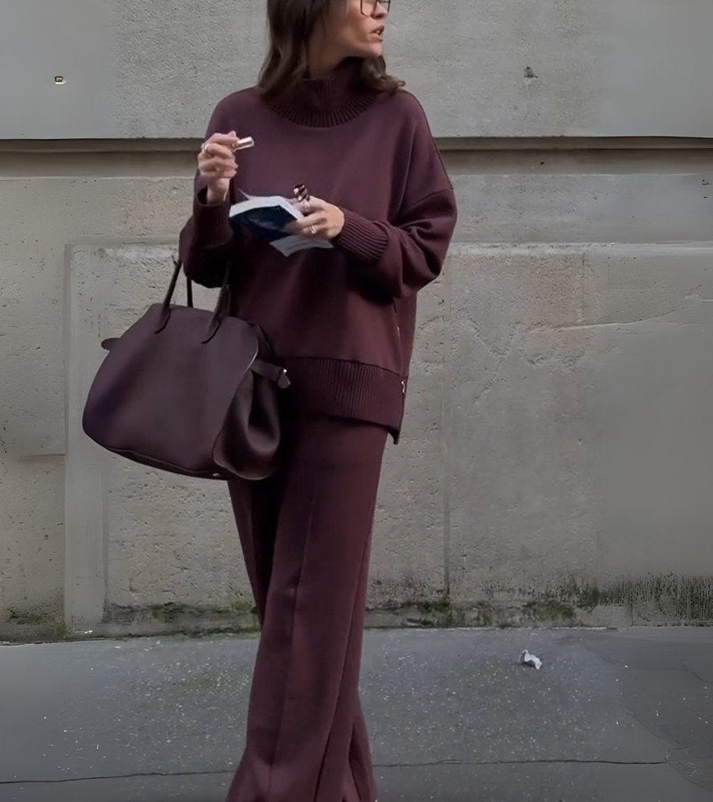 Woman in a burgundy two-piece set, standing outdoors with a large bag and reading material in hand.