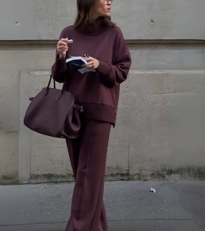 Woman in a burgundy two-piece set, standing outdoors with a large bag and reading material in hand.