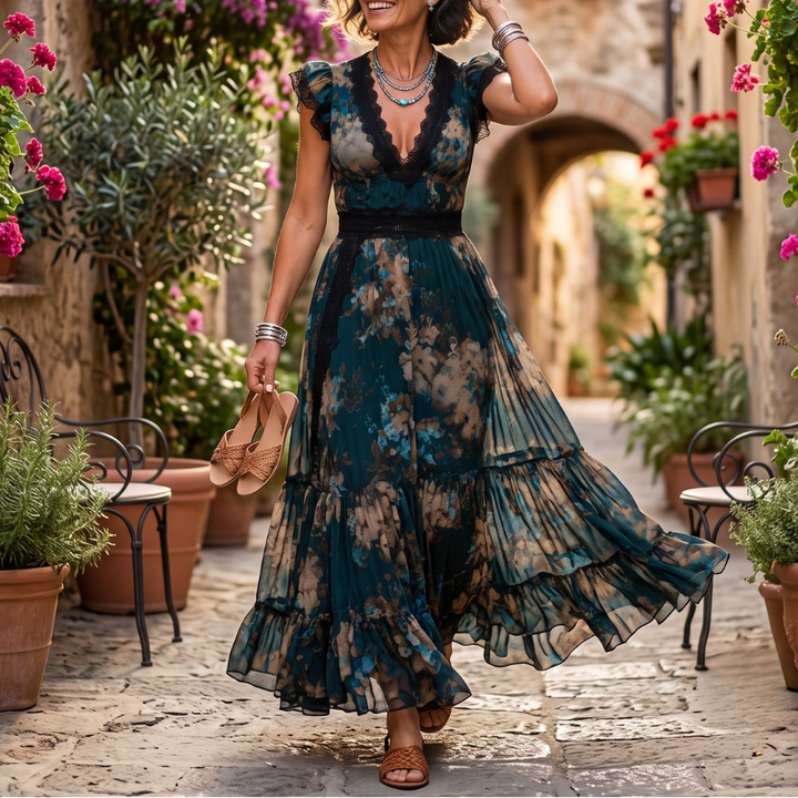 Flowy dark-patterned maxi dress with lace trim worn by a woman walking on a cobblestone street lined with vibrant flowers and potted plants.